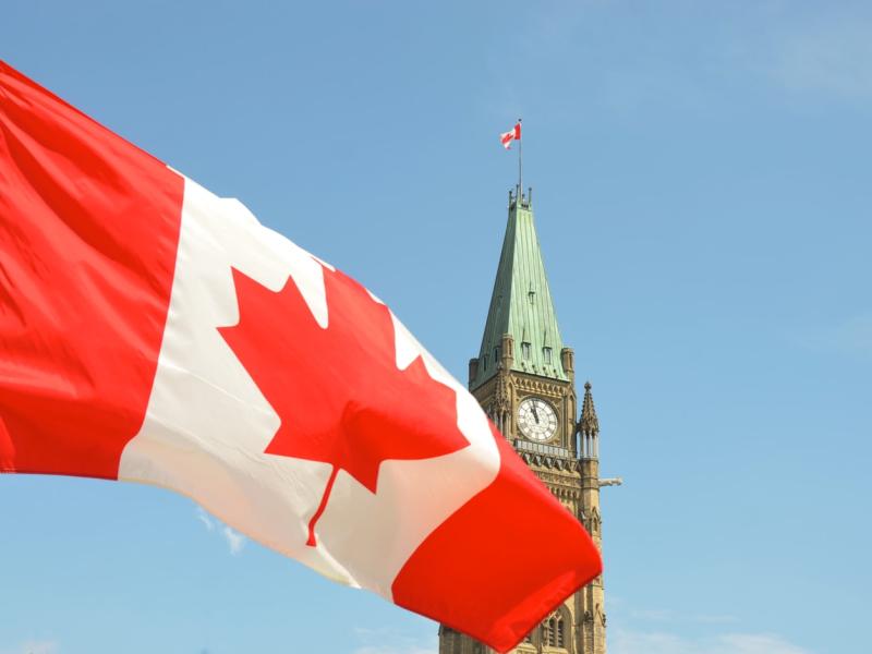 A Canadian flag waving in the foreground with the Peace Tower of the Canadian Parliament Buildings visible in the background against a blue sky.