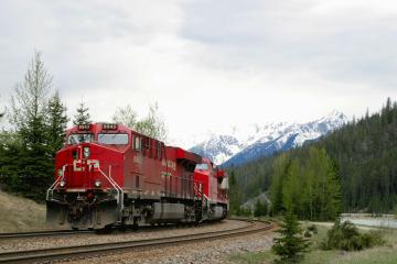 A red VIA Rail locomotive pulling a passenger train along a curved track through a mountainous landscape with snow-capped peaks and coniferous trees.