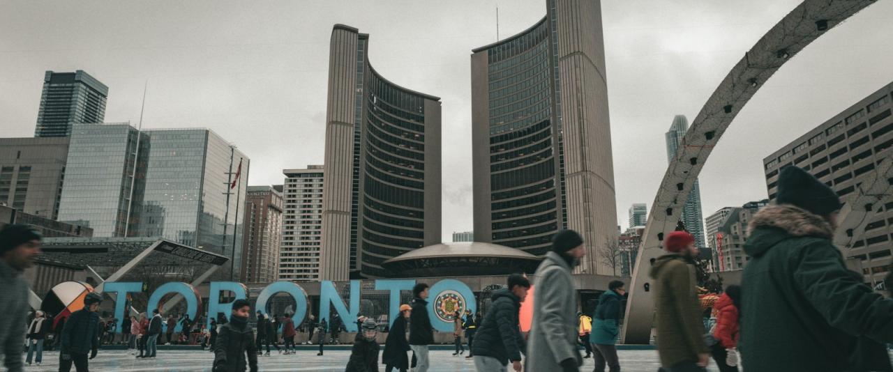 A group of people ice skating outdoors in a plaza with a large blue "TORONTO" sign and the distinctive curved Toronto City Hall buildings in the background.