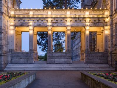 A stone colonnade with classical columns and a balustrade illuminated by string lights at dusk, featuring flower beds in the foreground and a view of the main building's facade through the arches.