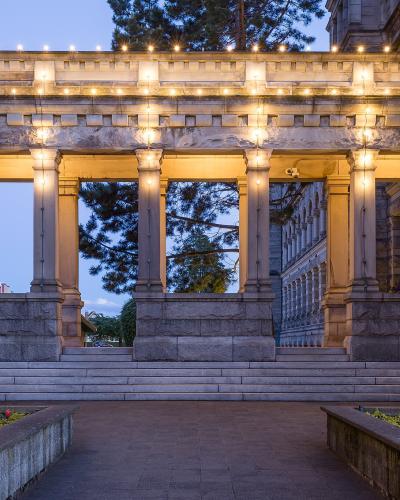 A stone colonnade with classical columns and a balustrade illuminated by string lights at dusk, featuring flower beds in the foreground and a view of the main building's facade through the arches.