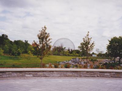 A large geodesic dome structure situated on a grassy hill surrounded by trees, viewed from a paved plaza with a low stone wall in the foreground.