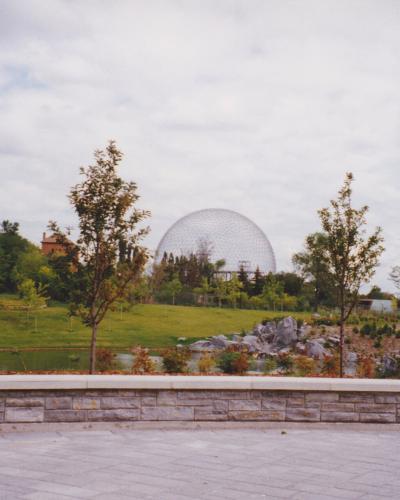 A large geodesic dome structure situated on a grassy hill surrounded by trees, viewed from a paved plaza with a low stone wall in the foreground.