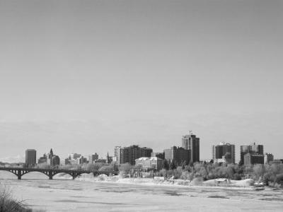 A black and white photograph showing a multi-arched bridge spanning a partially frozen river with a city skyline in the background.