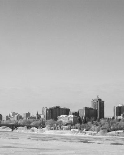 A black and white photograph showing a multi-arched bridge spanning a partially frozen river with a city skyline in the background.