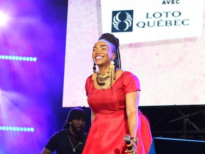 A woman in a red dress performs on stage with a guitarist behind her, in front of a large screen displaying the logo for "Festival Juste pour rire" and "Loto Québec".