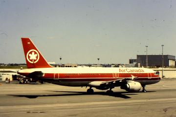 An Air Canada Airbus A320 aircraft is parked on the tarmac at an airport, with terminal buildings and light poles visible in the background.