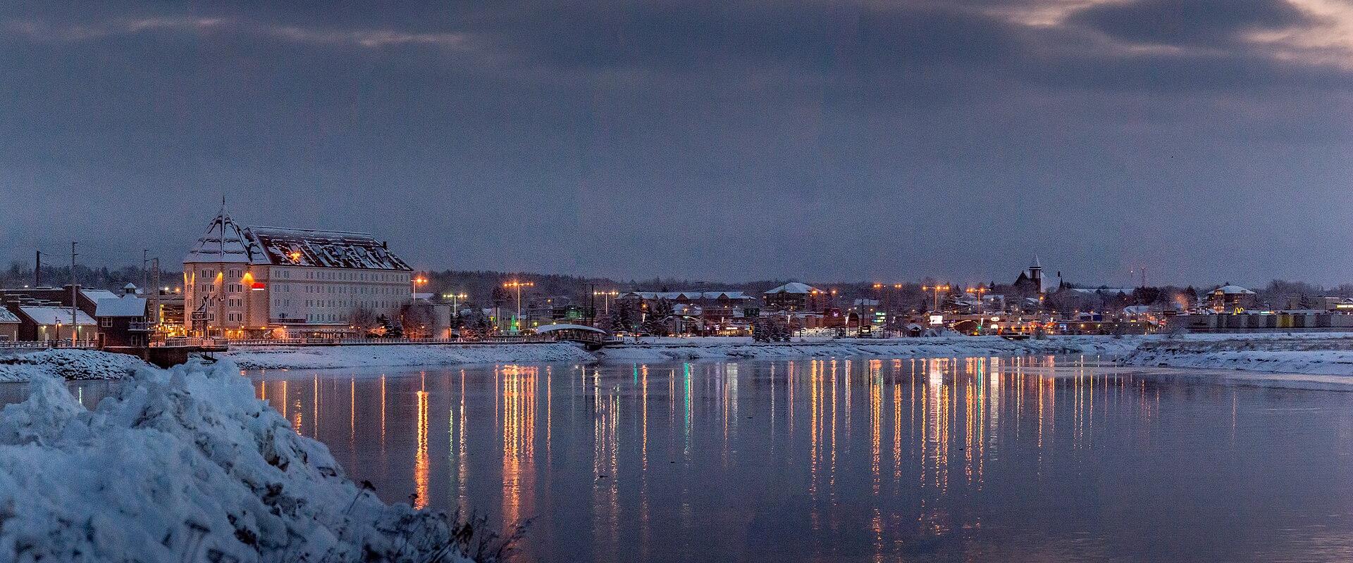 A winter evening view of a city skyline across a wide river, featuring a large white hotel with a distinctive peaked roof on the left bank and reflections of city lights on the water.