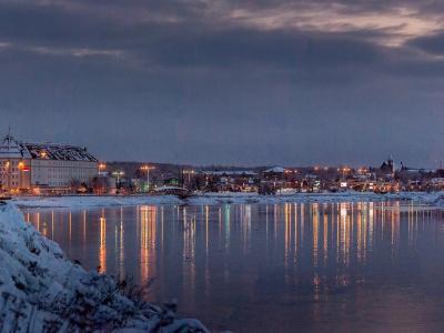 A winter evening view of a city skyline across a wide river, featuring a large white hotel with a distinctive peaked roof on the left bank and reflections of city lights on the water.