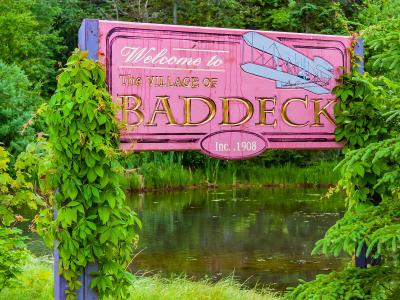 A large pink wooden welcome sign reading "Welcome to The VILLAGE OF BADDECK" with a biplane illustration and "Inc. 1908" text, framed by green foliage and situated next to a body of water.