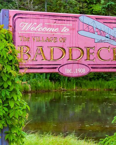 A large pink wooden welcome sign reading "Welcome to The VILLAGE OF BADDECK" with a biplane illustration and "Inc. 1908" text, framed by green foliage and situated next to a body of water.