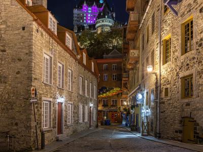 A cobblestone street in a historic district at night, flanked by old stone buildings, with the illuminated Château Frontenac visible on the cliff in the background.