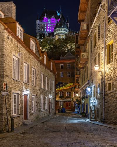 A cobblestone street in a historic district at night, flanked by old stone buildings, with the illuminated Château Frontenac visible on the cliff in the background.