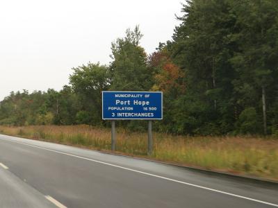 A blue rectangular highway welcome sign reading "MUNICIPALITY OF Port Hope" situated on the grassy shoulder of a multi-lane highway, with trees and overcast sky in the background.