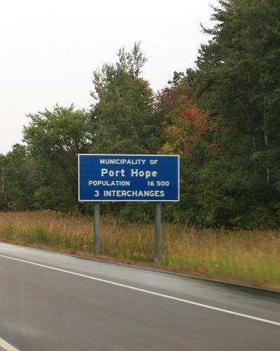 A blue rectangular highway welcome sign reading "MUNICIPALITY OF Port Hope" situated on the grassy shoulder of a multi-lane highway, with trees and overcast sky in the background.
