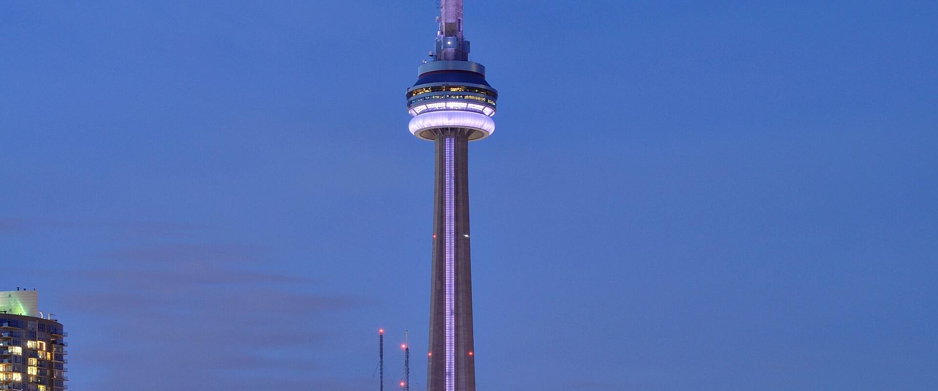 A night photograph of the CN Tower illuminated in purple and white light, dominating the Toronto skyline with surrounding skyscrapers and the waterfront in the foreground.