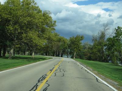 A paved two-lane road with yellow center lines curving through a grassy, tree-lined park area, with water visible to the right and overcast skies above.