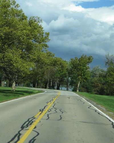 A paved two-lane road with yellow center lines curving through a grassy, tree-lined park area, with water visible to the right and overcast skies above.