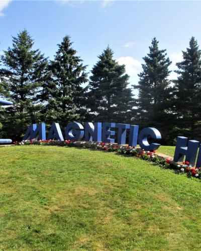 A large outdoor sign spelling "MAGNETIC HILL" in blue block letters with a red and blue horseshoe magnet sculpture to the left, set on a grassy slope with evergreen trees in the background.