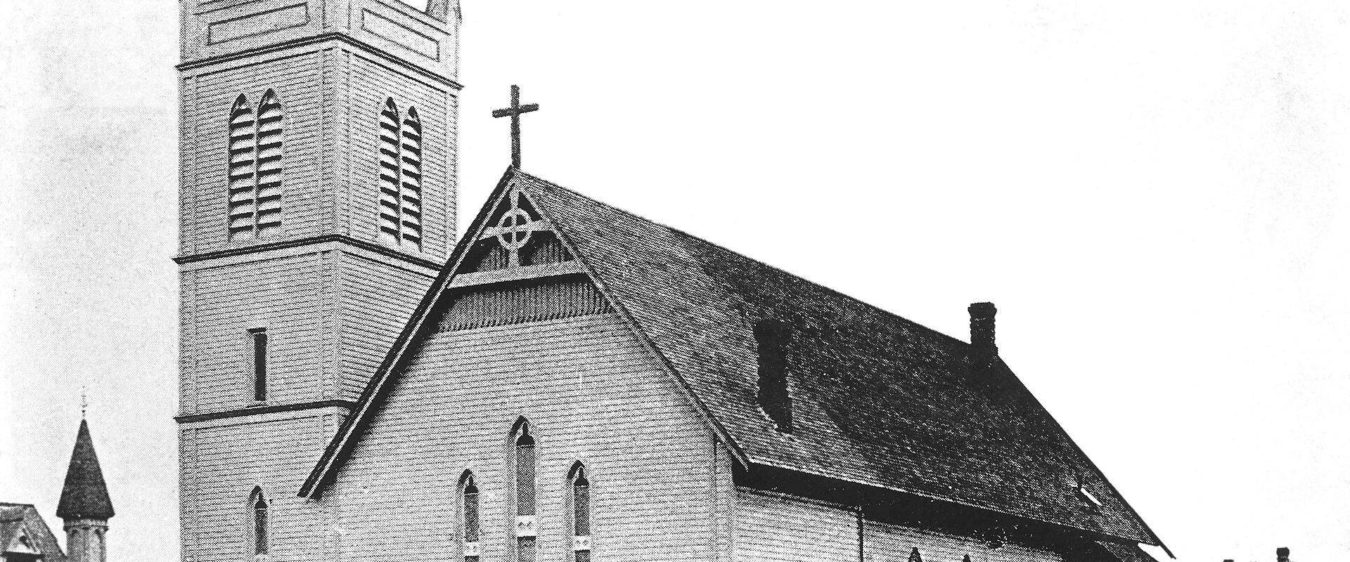 A black and white historical photograph of a wooden church building featuring a tall, square bell tower with louvered openings and a steep gabled roof.