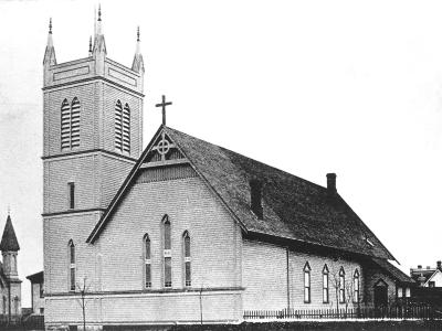 A black and white historical photograph of a wooden church building featuring a tall, square bell tower with louvered openings and a steep gabled roof.