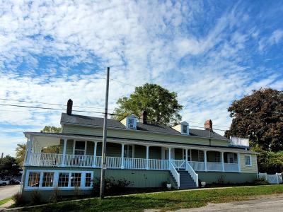 A two-story red brick house with white window frames, dark shutters, and a manicured garden featuring a conical evergreen tree and a small informational sign in the front yard.