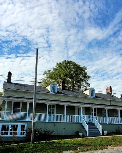 A two-story red brick house with white window frames, dark shutters, and a manicured garden featuring a conical evergreen tree and a small informational sign in the front yard.