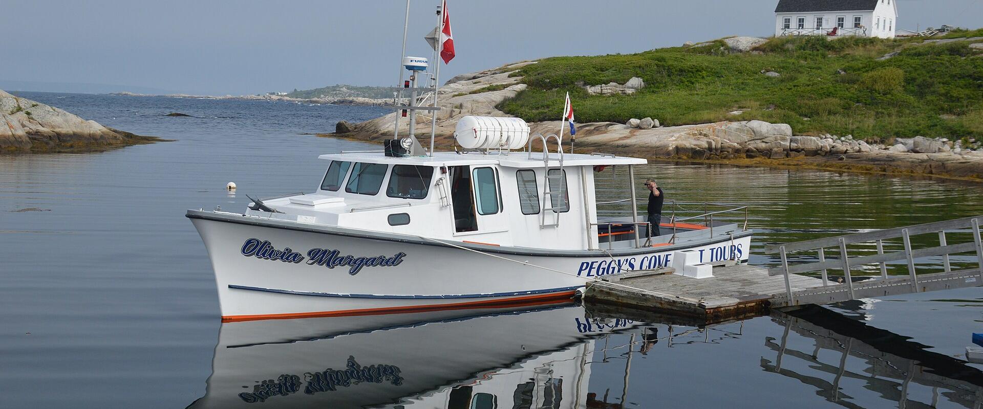 A white tour boat named 'Olivia Margaret' is docked at a wooden pier in a calm harbor, with a white house on a grassy hill in the background.
