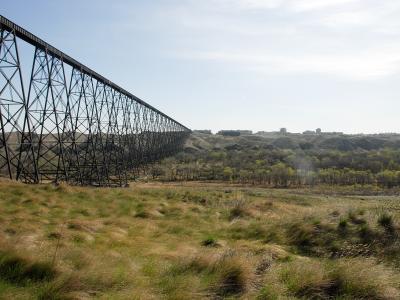 A large steel trestle bridge spanning a deep, grassy river valley with a city skyline visible on the distant horizon.