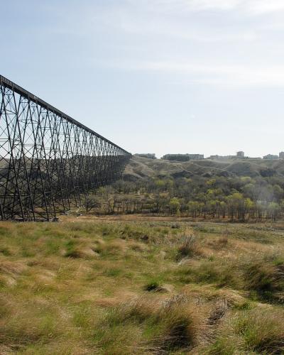 A large steel trestle bridge spanning a deep, grassy river valley with a city skyline visible on the distant horizon.