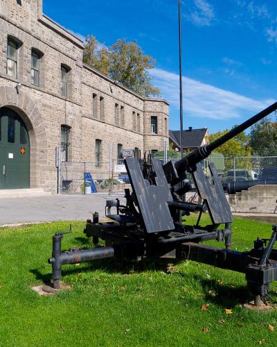 A large black anti-aircraft gun mounted on a grassy lawn in front of a two-story stone building with an arched green door and multiple windows.