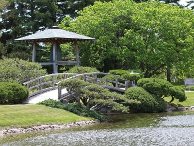A wooden arched bridge crossing a pond, with a traditional Japanese-style wooden pavilion (gazebo) visible in the background, surrounded by manicured trees and shrubbery.