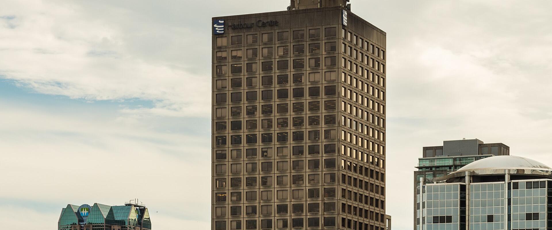 A tall, rectangular office building topped with a distinctive circular observation deck and a flagpole flying the Canadian flag, set against a cloudy sky with other city buildings in the foreground and background.