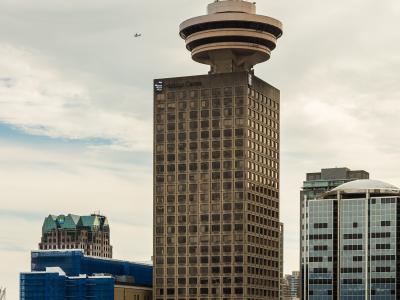 A tall, rectangular office building topped with a distinctive circular observation deck and a flagpole flying the Canadian flag, set against a cloudy sky with other city buildings in the foreground and background.