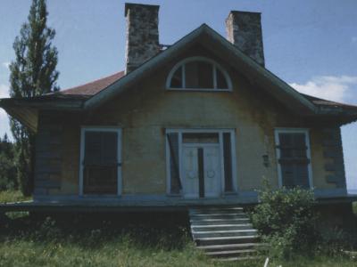 A yellow, single-story wooden house with two stone chimneys, a central white door, and a semi-circular window above it, situated on a grassy slope near water.