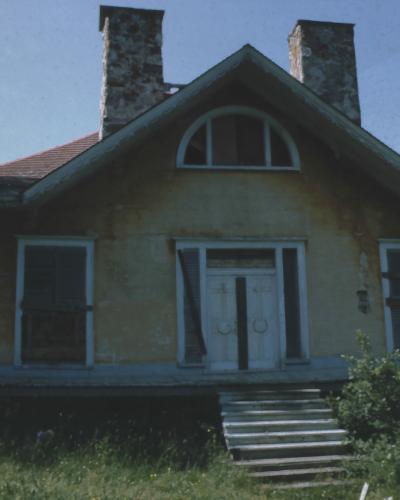 A yellow, single-story wooden house with two stone chimneys, a central white door, and a semi-circular window above it, situated on a grassy slope near water.
