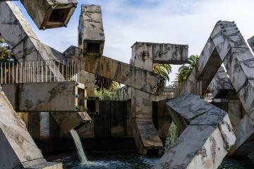 A large, brutalist concrete sculpture featuring interlocking geometric blocks and waterfalls, set in an outdoor plaza with palm trees visible in the background.