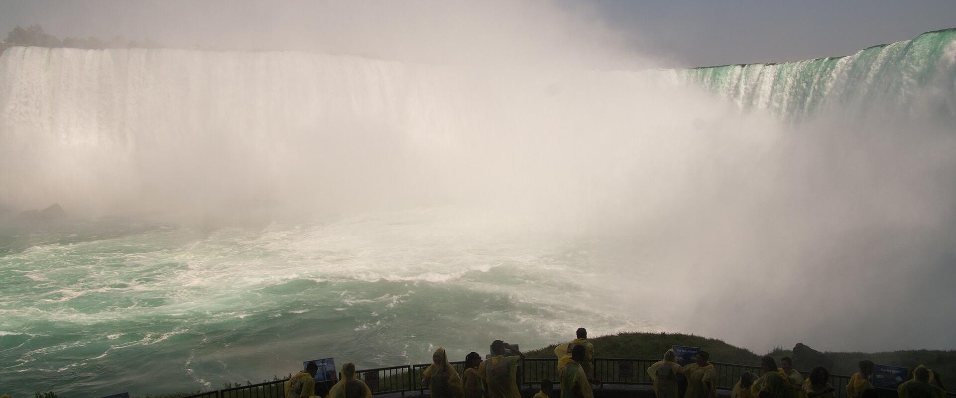 A panoramic view from an outdoor observation deck showing a massive waterfall with a large crowd of people wearing yellow rain ponchos.