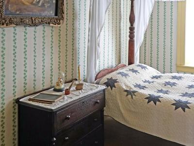 An interior bedroom scene featuring a four-poster bed with a star-patterned quilt, a dark wooden dresser, and walls covered in green and white striped wallpaper with a floral border.