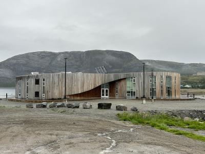 A modern, curved wooden building with large windows and a distinctive undulating roofline, situated on a gravel lot near water with a large mountain in the background.