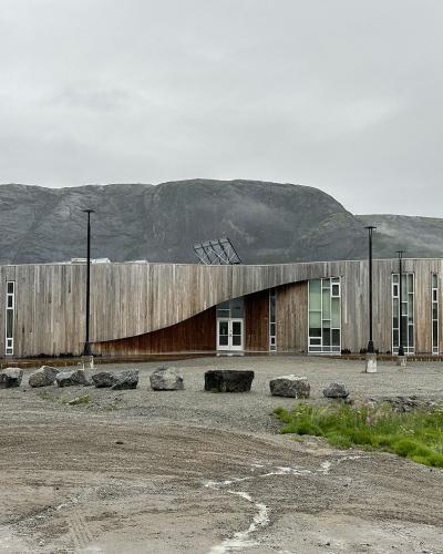 A modern, curved wooden building with large windows and a distinctive undulating roofline, situated on a gravel lot near water with a large mountain in the background.