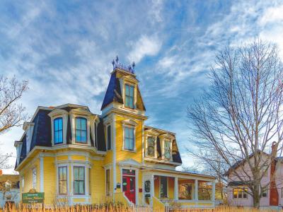 A yellow Victorian-style house with a prominent tower and mansard roof, featuring a green sign in the front yard that reads "Thomas Williams House".