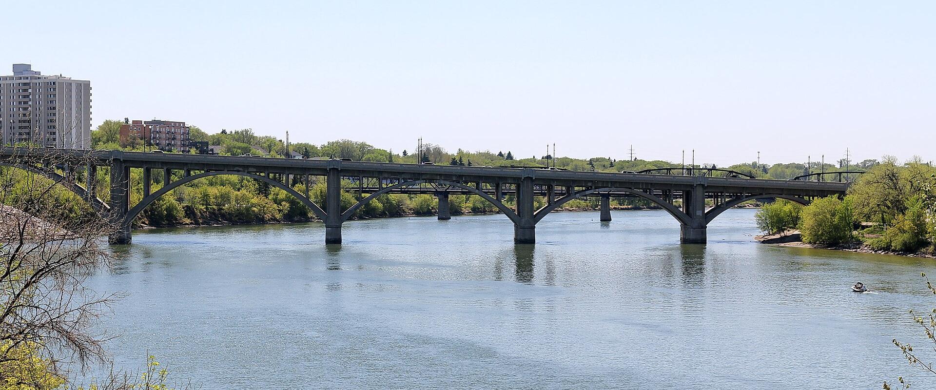 A concrete arch bridge spanning a wide river with urban buildings visible on the left bank and trees lining the riverbanks.