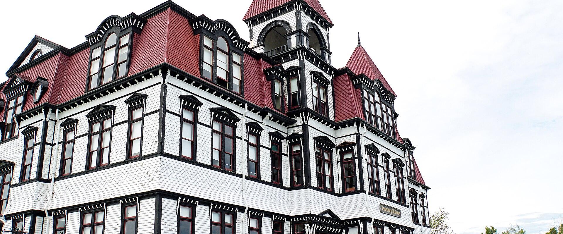 A low-angle photograph of a large, multi-story wooden building with white siding, black trim, and a red mansard roof, featuring a prominent corner tower and arched windows.