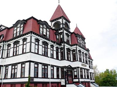 A low-angle photograph of a large, multi-story wooden building with white siding, black trim, and a red mansard roof, featuring a prominent corner tower and arched windows.