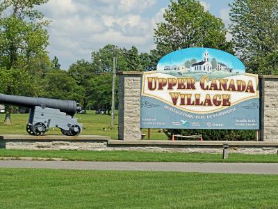 A large entrance sign for "Upper Canada Village Heritage Park" featuring a painted church and a cannon on display to the left.