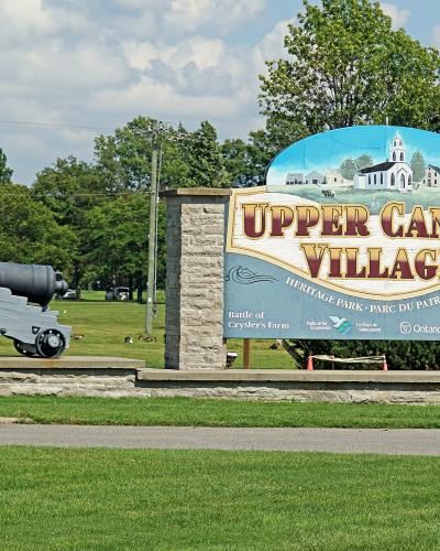 A large entrance sign for "Upper Canada Village Heritage Park" featuring a painted church and a cannon on display to the left.
