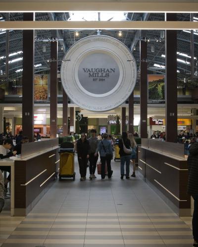 The image shows the interior of a shopping mall food court area, featuring a large circular hanging sign with the text "VAUGHAN MILLS EST 2004" in the center.