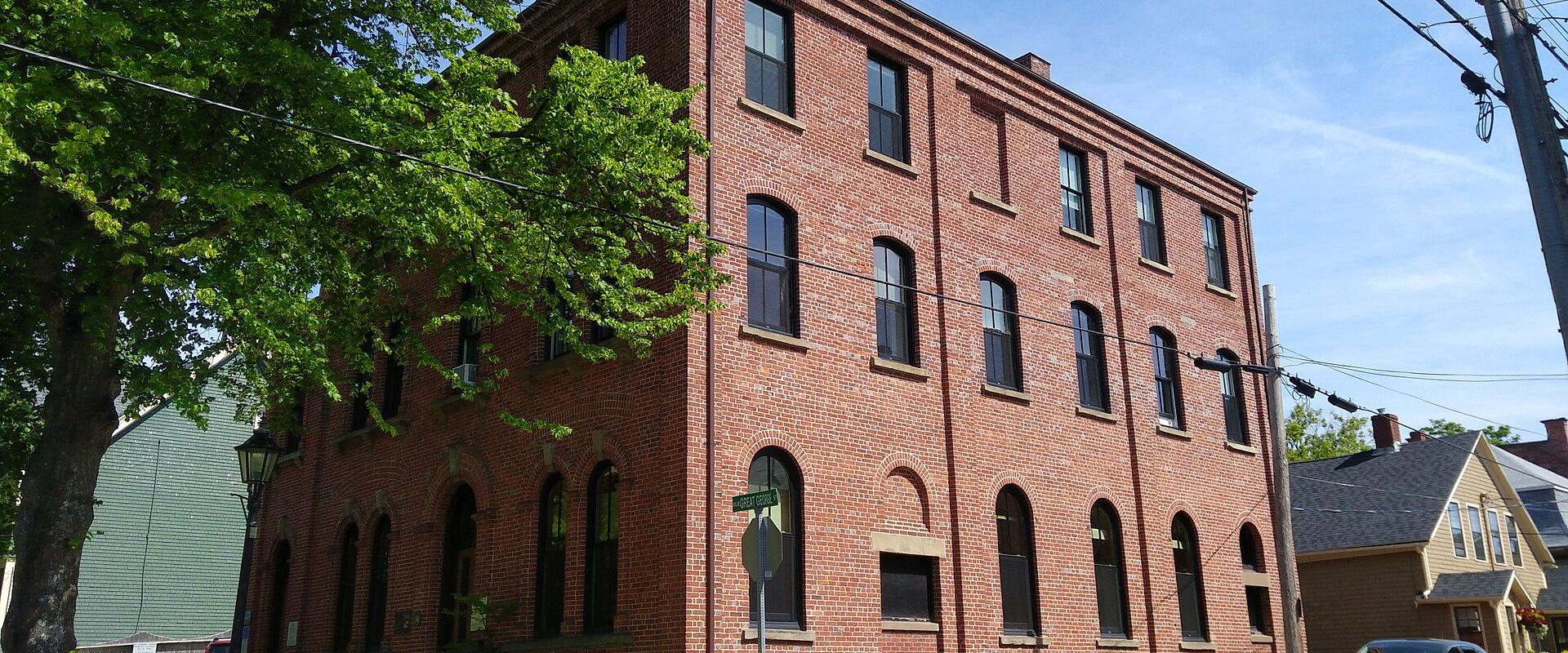 A three-story red brick building with arched windows on the ground floor and rectangular windows above, situated on a street corner with a green street sign reading "Fitzroy St" and a car parked nearby.