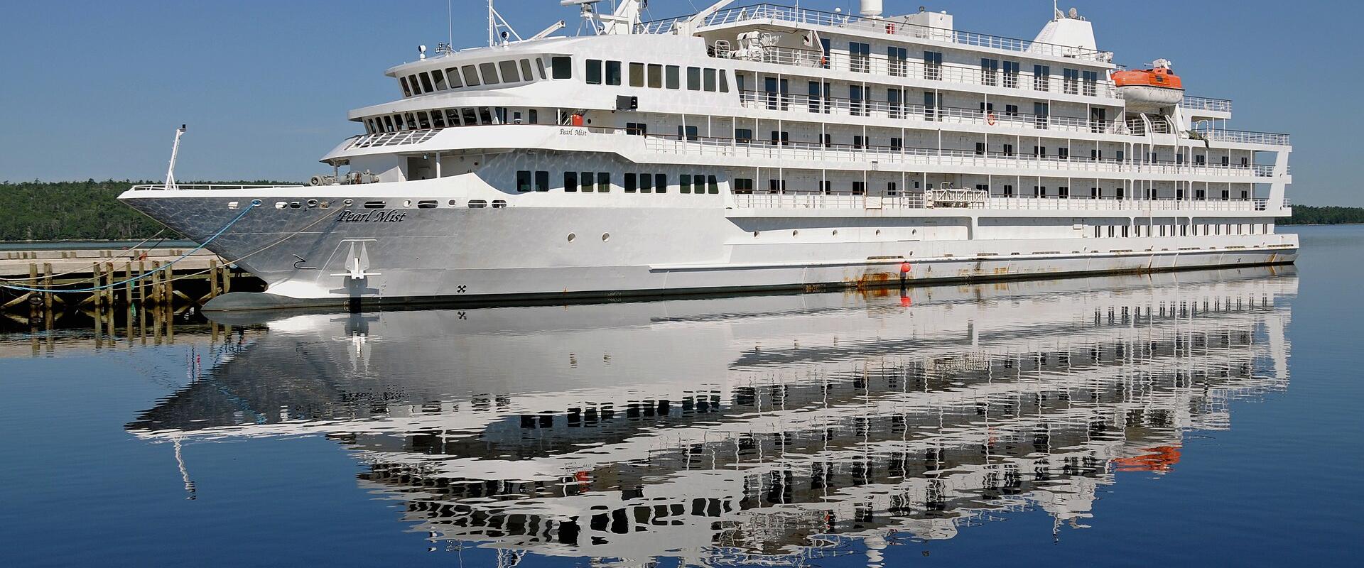 A large white cruise ship named "Pearl Mist" is docked at a wooden pier in calm water with a forested shoreline in the background.